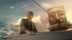 A man in a suit sits in a boat next to a large painting of a face against a cloudy sky.