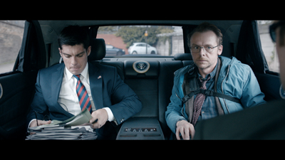 A man in a suit looks at papers while another man in a blue jacket and glasses looks forward in a car backseat.