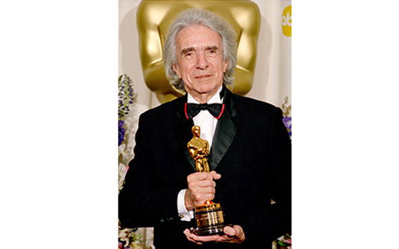 A man in a black tuxedo holding a gold Oscar award in front of a large gold Oscar silhouette.