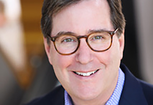 A smiling man with brown hair and brown-rimmed glasses wearing a blue blazer and checkered shirt.