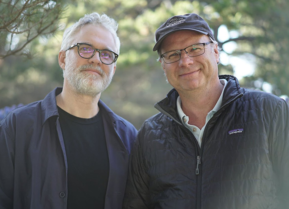 Two men with glasses smiling while standing in front of trees.