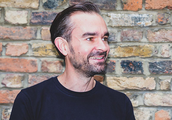 A man with short dark hair and a beard smiling while looking to the side against a brick wall.