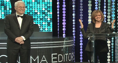 A man in a tuxedo and a woman in a black blazer stand on a stage with a blue and white patterned backdrop.