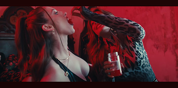 Two women in a room with red lighting; one eats a cherry while the other holds a bottle of red liquid.