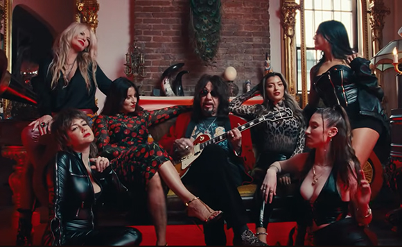 A group of seven musicians in dark, stylish clothing posing in a room with red lighting and brick walls.