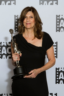 A woman with brown hair wearing a black dress holds a silver trophy against a white patterned background.