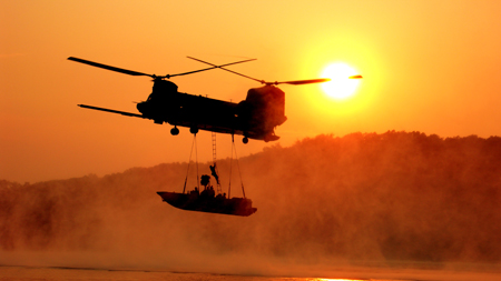 Silhouette of a dual-rotor helicopter transporting a small boat against a bright setting sun.
