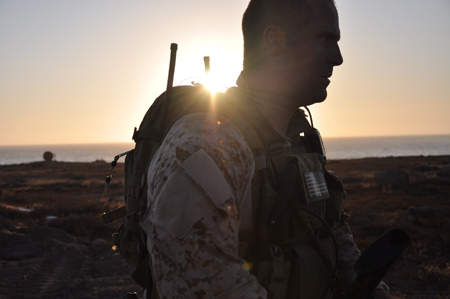 Silhouette of a soldier in camouflage gear standing in a field during sunset.