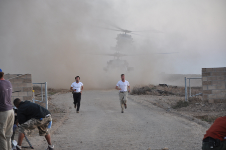 Two men in white shirts run down a dirt path toward the camera amidst dust from landing helicopters.