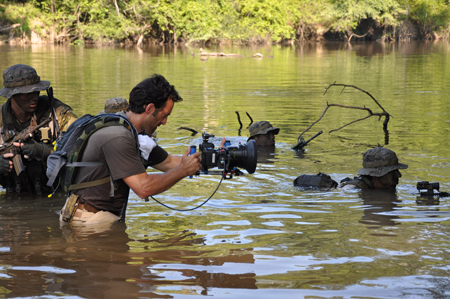 A man in a brown shirt holds a professional camera while wading through a river alongside others in camouflage.