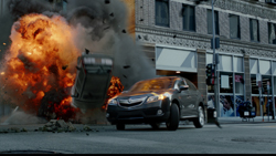 A dark car drives past a massive orange and black explosion on a city street.
