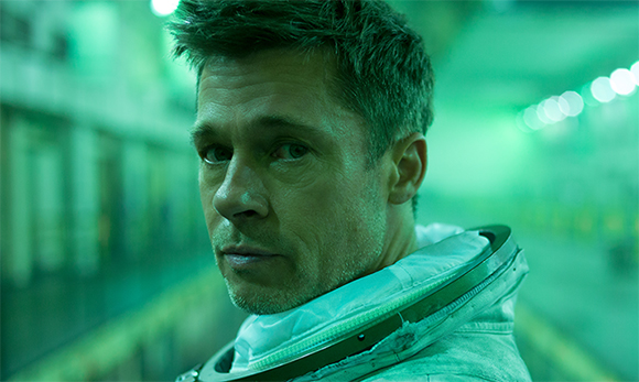 A man with short brown hair wearing a white spacesuit looks intensely toward the camera against a green background.