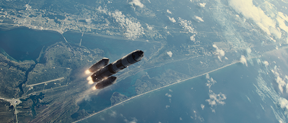 A multi-stage rocket launches upward over a coastal landscape with blue water and scattered clouds.
