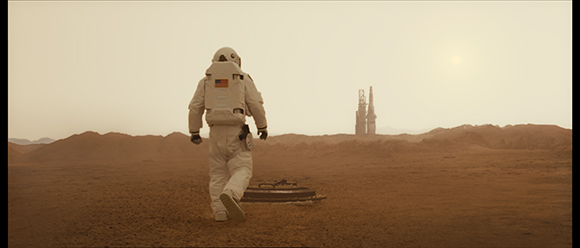 An astronaut in a white spacesuit walks across a vast, reddish desert toward distant towers under a hazy sky.