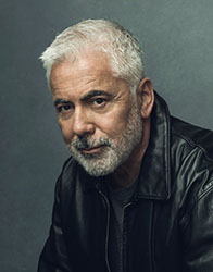 A man with short gray hair and a gray beard wearing a black leather jacket against a gray background.