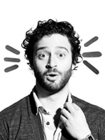 Black and white portrait of a man with curly hair and a beard looking surprised with hand near his collar.