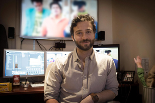 A man with dark hair and a beard sits in an office with multiple computer monitors in the background.