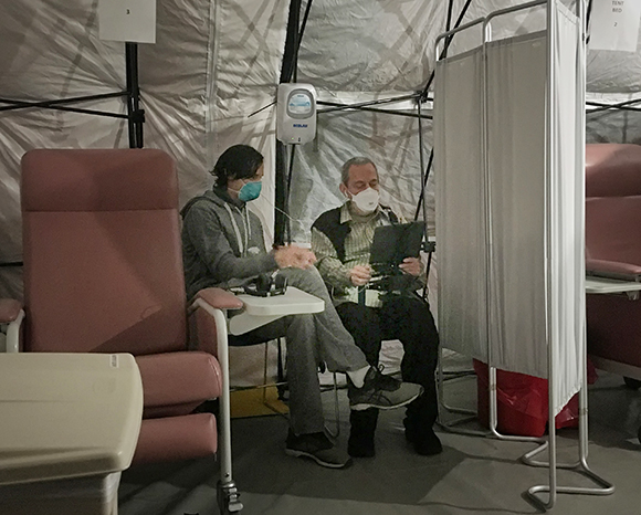 Two people in masks sit in chairs inside a white medical tent with privacy screens.