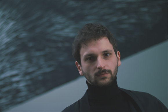 A man with dark hair and a beard wearing a black turtleneck against a dark, textured background.