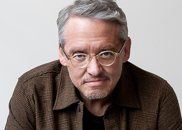 A man with gray hair and round glasses wearing a brown textured jacket looking at the camera.