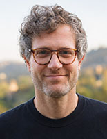 A man with curly hair and glasses smiling against a blurred outdoor background.