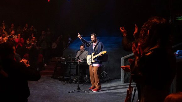 A man in a blue hoodie sings and points upward on a dimly lit stage while a keyboardist plays behind him.