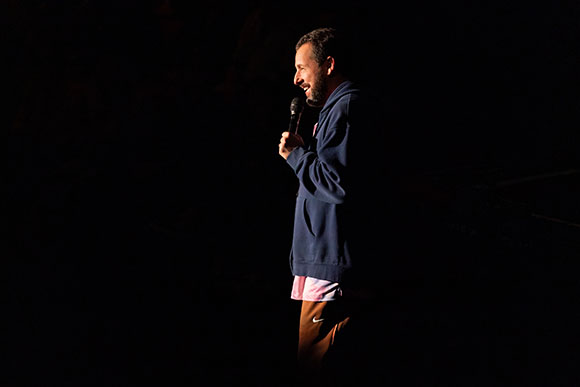 A man in a blue hoodie holds a microphone and speaks against a black background.