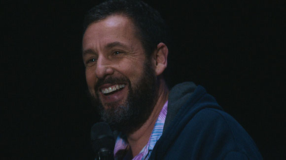 A man with a dark beard and short hair smiles while wearing a blue hoodie and patterned shirt.