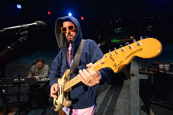 A man wearing orange sunglasses and a blue hoodie plays a Fender Stratocaster guitar on a dark stage.
