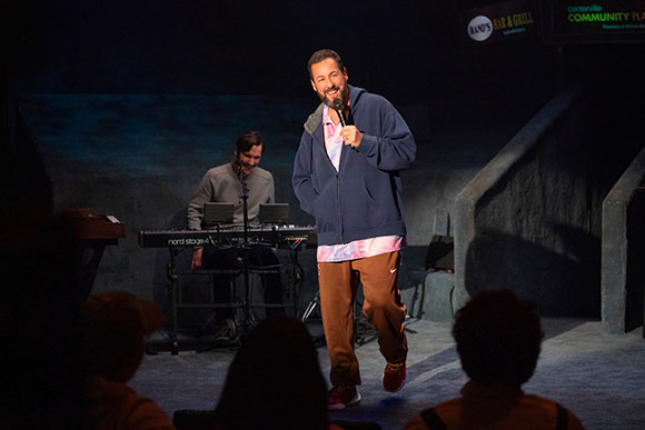 A man in a blue hoodie holds a microphone on a stage while a musician plays a keyboard behind him.
