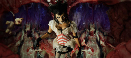A woman in a white ruffled corset and black gloves stands inside a dark, fleshy, organic-looking cavern.