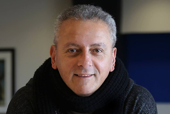 A middle-aged man with short graying hair and a black scarf smiling at the camera.