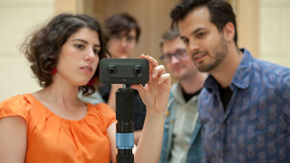 A group of young adults looking intently at a small black camera mounted on a tripod.