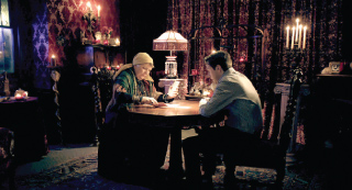 A person in a headscarf reads tarot cards for a seated man in a dark, candlelit room.