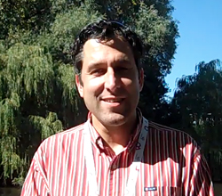A man with dark curly hair wearing a red and white striped shirt, smiling outdoors with trees in the background.