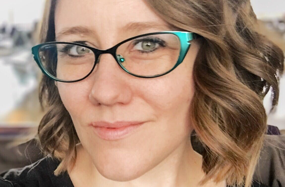 A woman with wavy brown hair and teal-rimmed glasses looking into the camera.