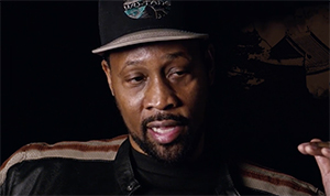 A man with a goatee wearing a black baseball cap and a black and brown striped jacket.