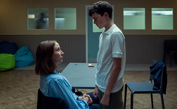 A woman in a blue shirt sits at a table looking up at a boy in a white polo shirt standing in a dim room.