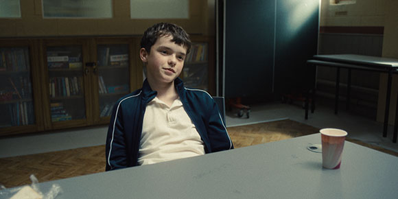A young boy in a white shirt and dark jacket sits at a grey table in a room with bookshelves.