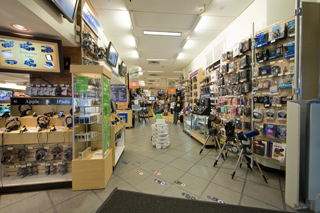 A long aisle in an electronics store filled with shelves of gadgets, headphones, and cameras.
