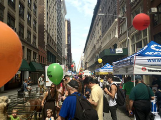 A crowded city street with tall buildings, event tents, and colorful balloons floating above the crowd.