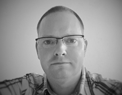 Black and white headshot of a man with short hair and glasses wearing a plaid shirt.