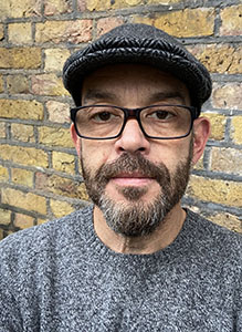 A man with a salt-and-pepper beard, glasses, and a dark flat cap stands in front of a brick wall.