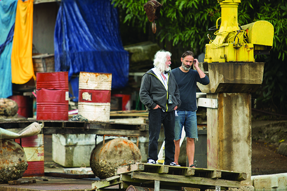 Two men stand near a large yellow crane and stacked barrels in an outdoor industrial setting.