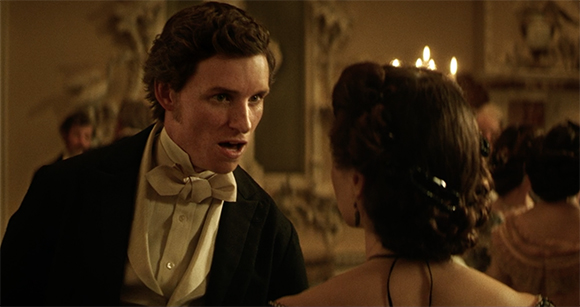 A man in a black tuxedo and white bow tie speaks to a woman with dark hair in a dimly lit ballroom.