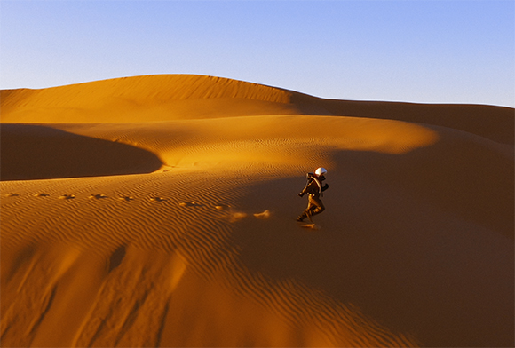 A person walking along the ridge of a large, sunlit sand dune under a clear sky.
