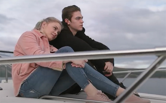 A young woman in a pink jacket leans her head on a young man's shoulder as they sit on a boat.