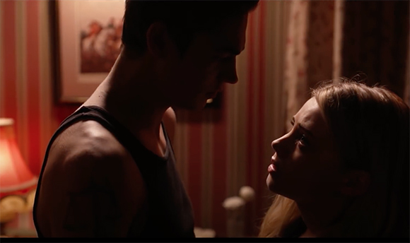 A man in a tank top looks down at a woman who looks up at him in a dark, warm-toned room.