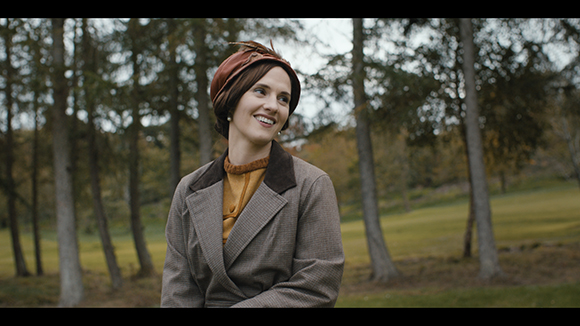 A smiling woman wearing a brown hat and a patterned blazer stands in a forest setting.