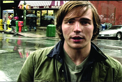 A man with light brown hair wearing a green jacket stands on a wet urban street.